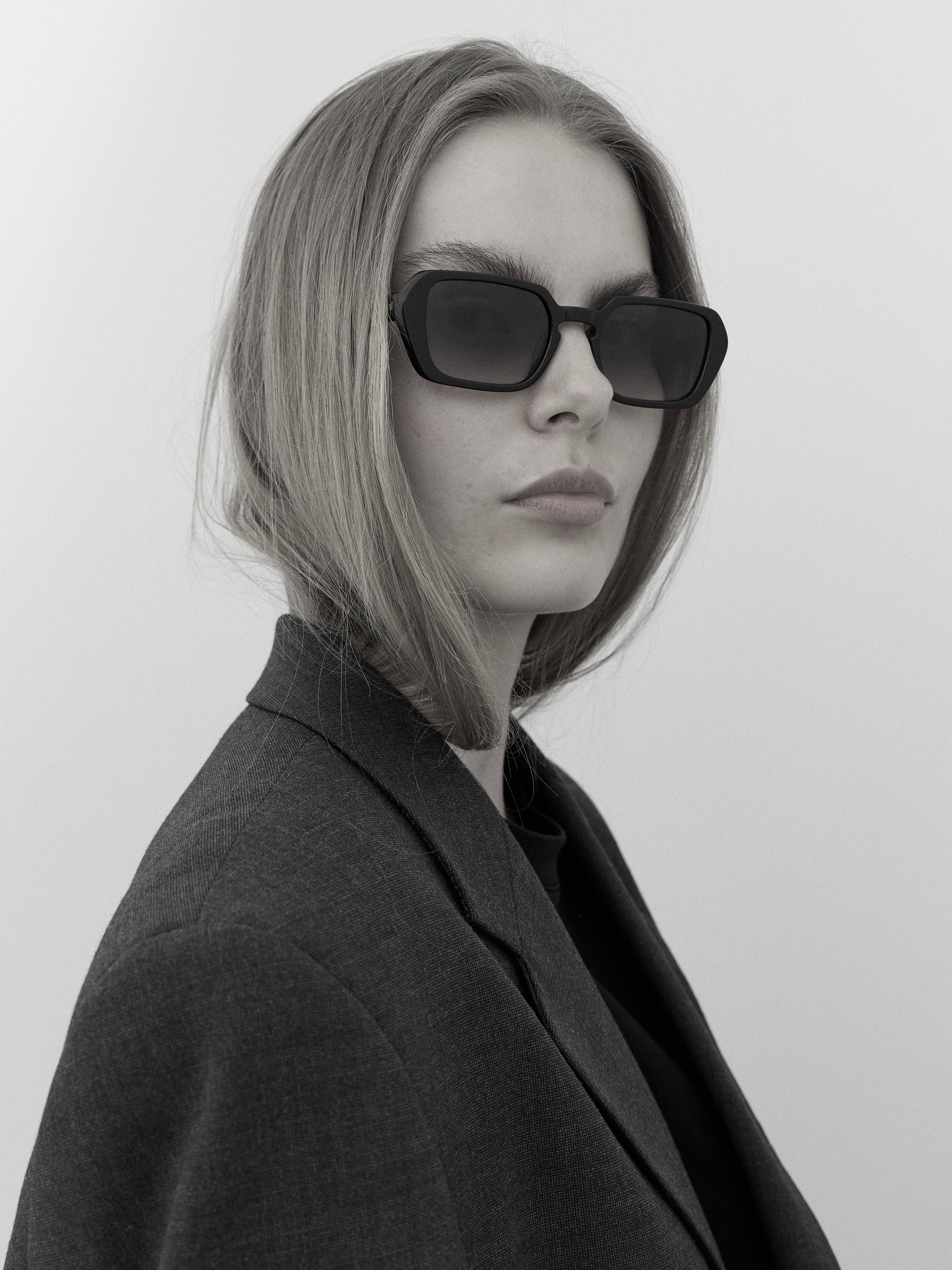 Female model wearing Marselis solid black sunglasses from Anatolí, styled with a charcoal blazer in a minimalist setting.