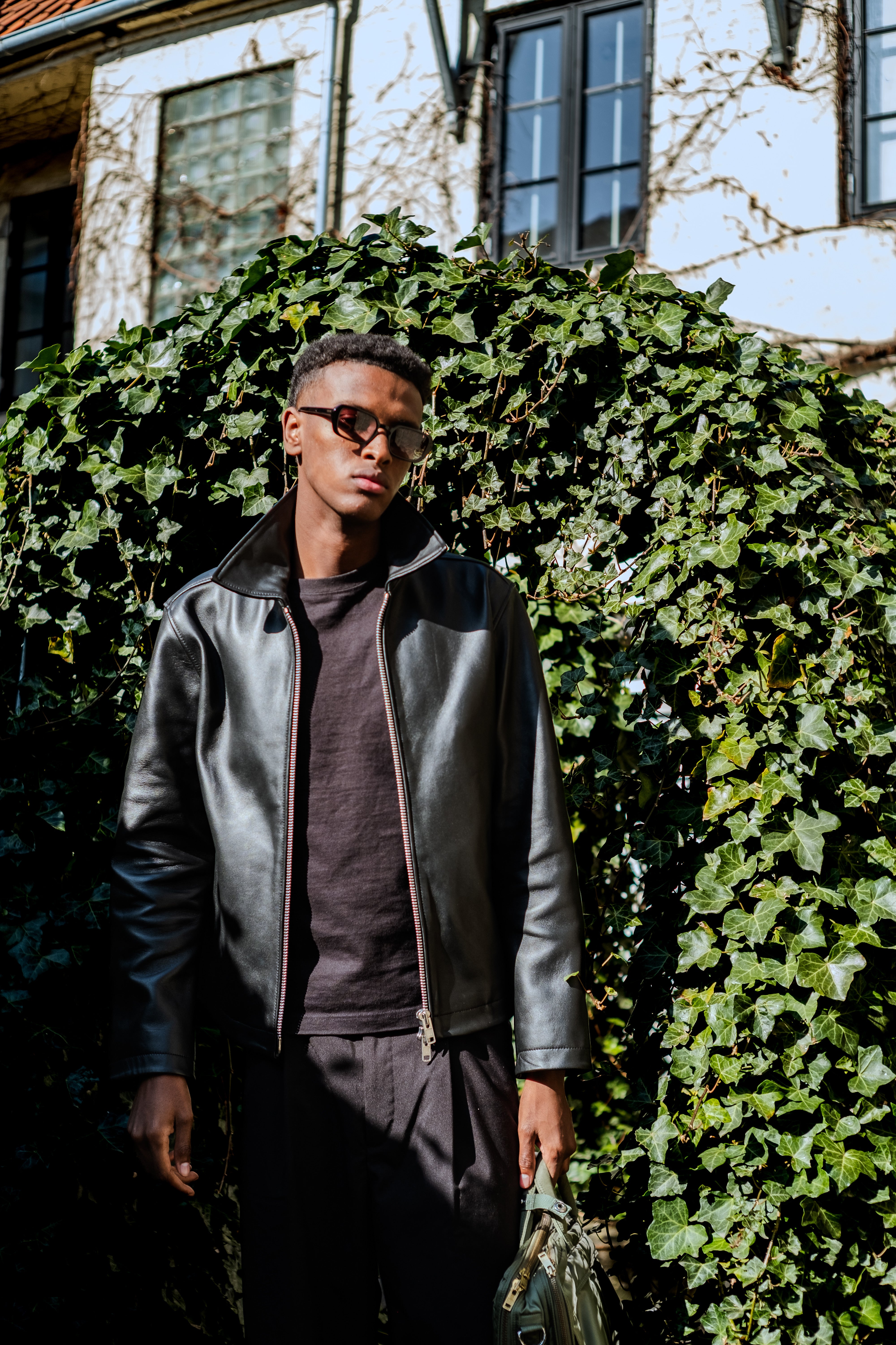 Full-body portrait of male model in leather jacket and tortoise frame sunglasses by Anatolí, standing in front of ivy-covered wall