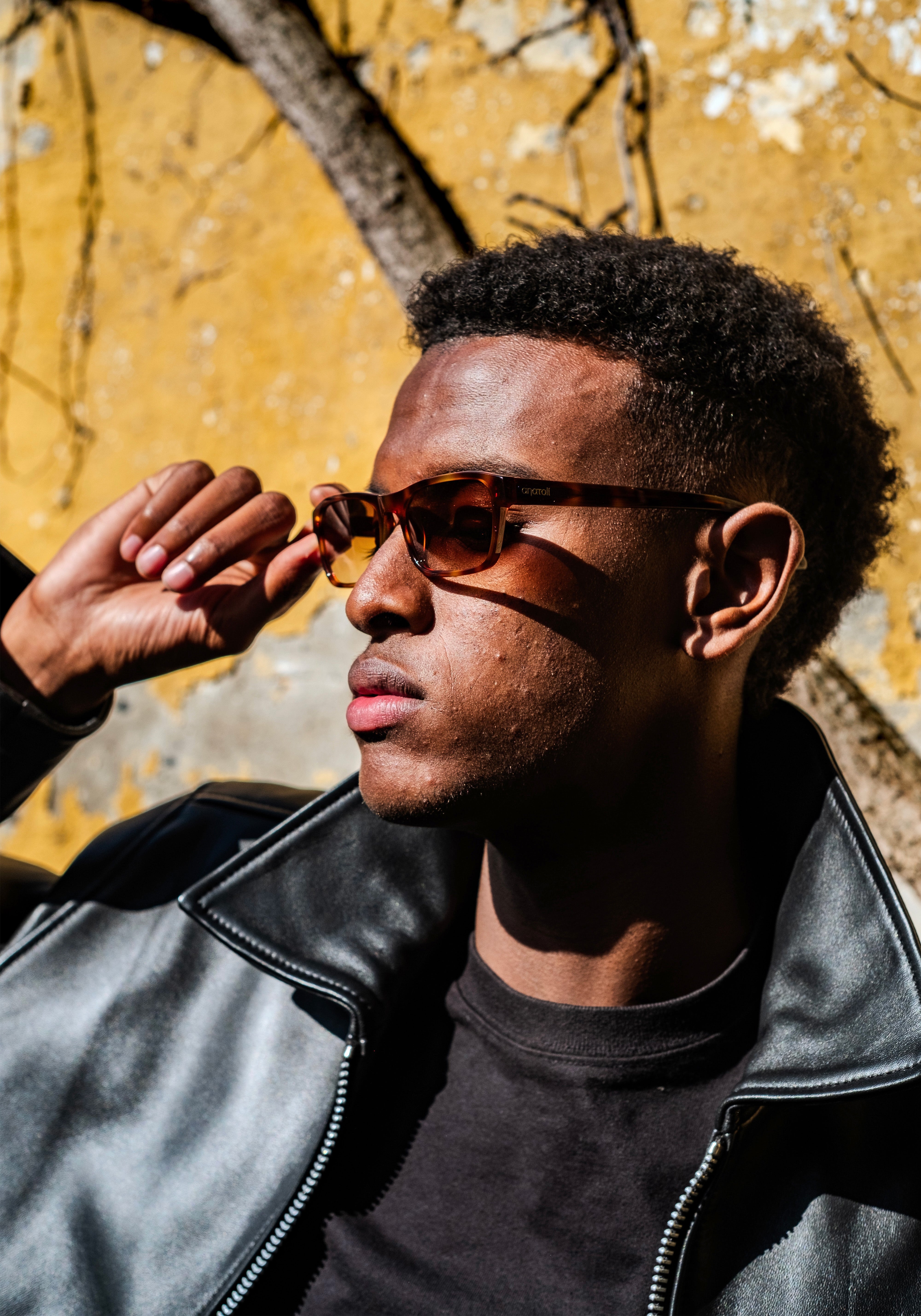 Male model wearing dark rectangular sunglasses from Anatolí, photographed in natural sunlight against a textured yellow wall.