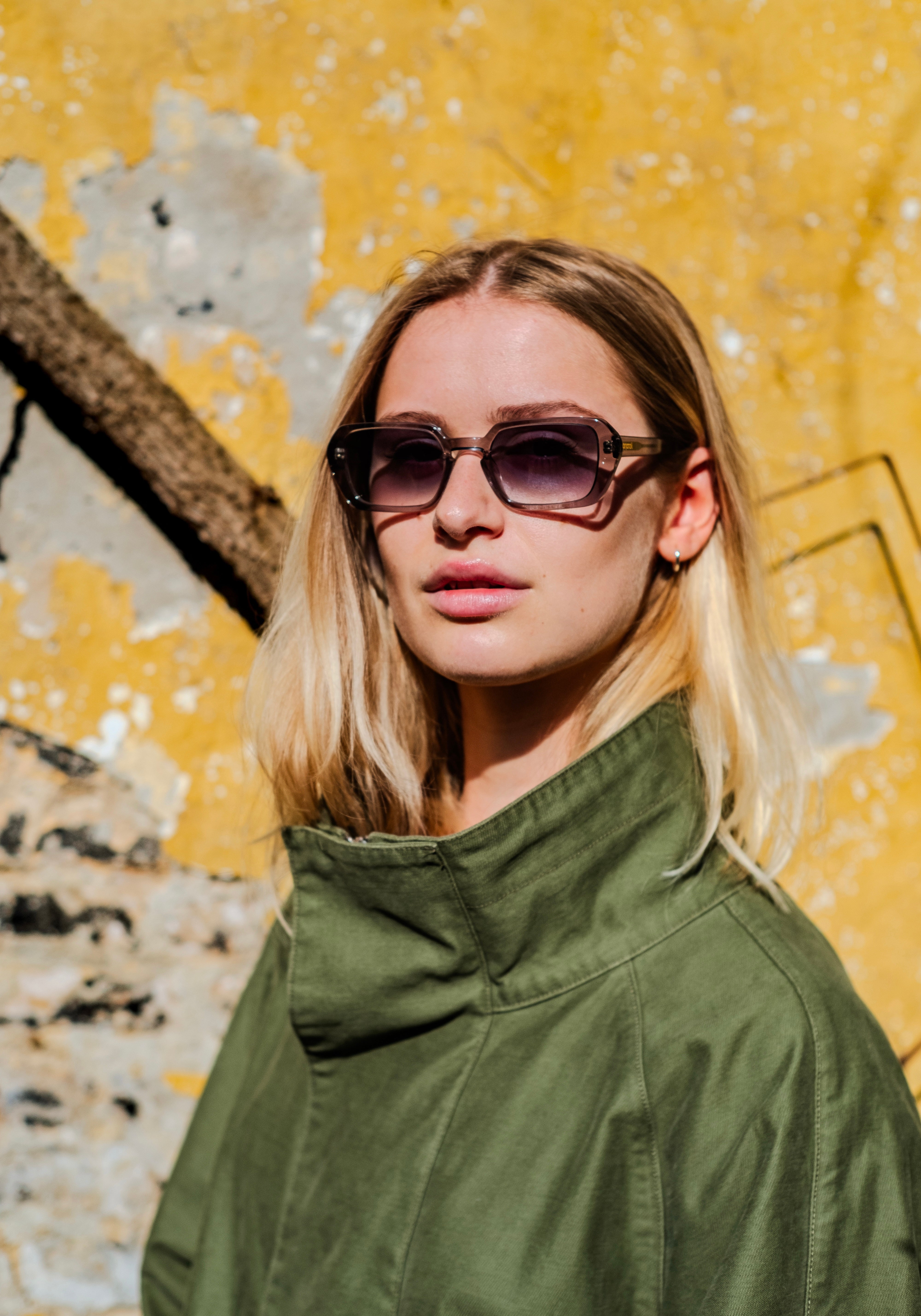Male model wearing dark rectangular sunglasses from Anatolí, photographed in natural sunlight against a textured yellow wall.